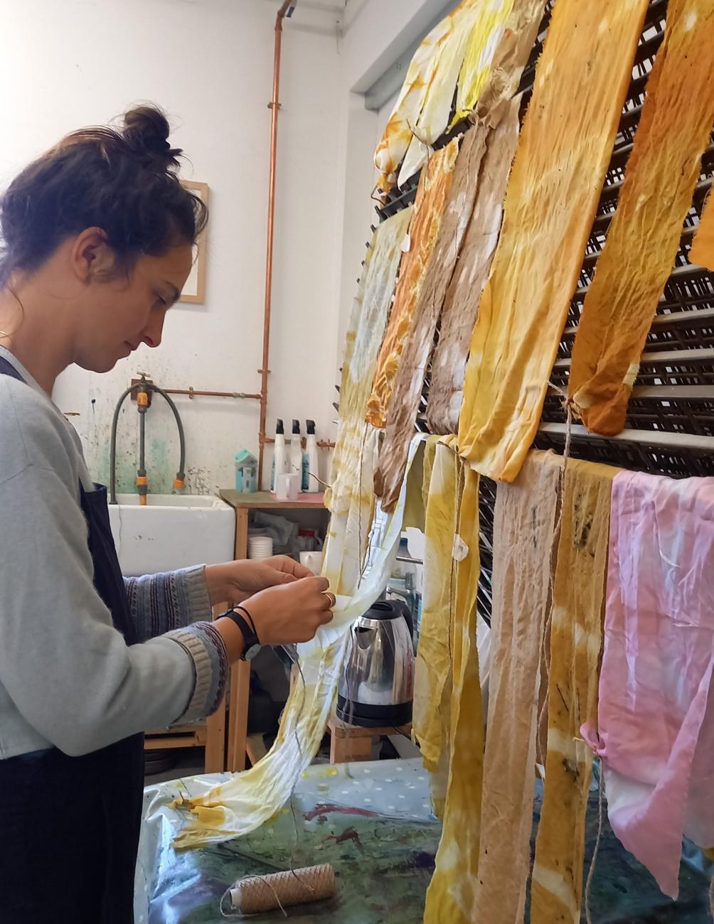Naturally dyed strips of fabric are hanging up to dry. A person is holding the end of one of the lengths of fabric. The colours are lovely soft yellows, oranges and browns.