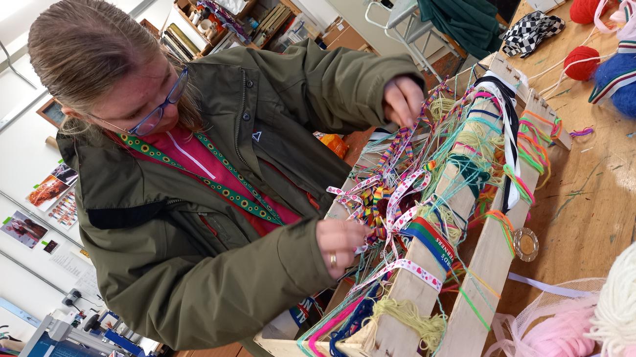 A person leans over a wooden frame, weaving brightly coloured wool, laces and threads back and forth and in and out to make a mesh of colour and texture.