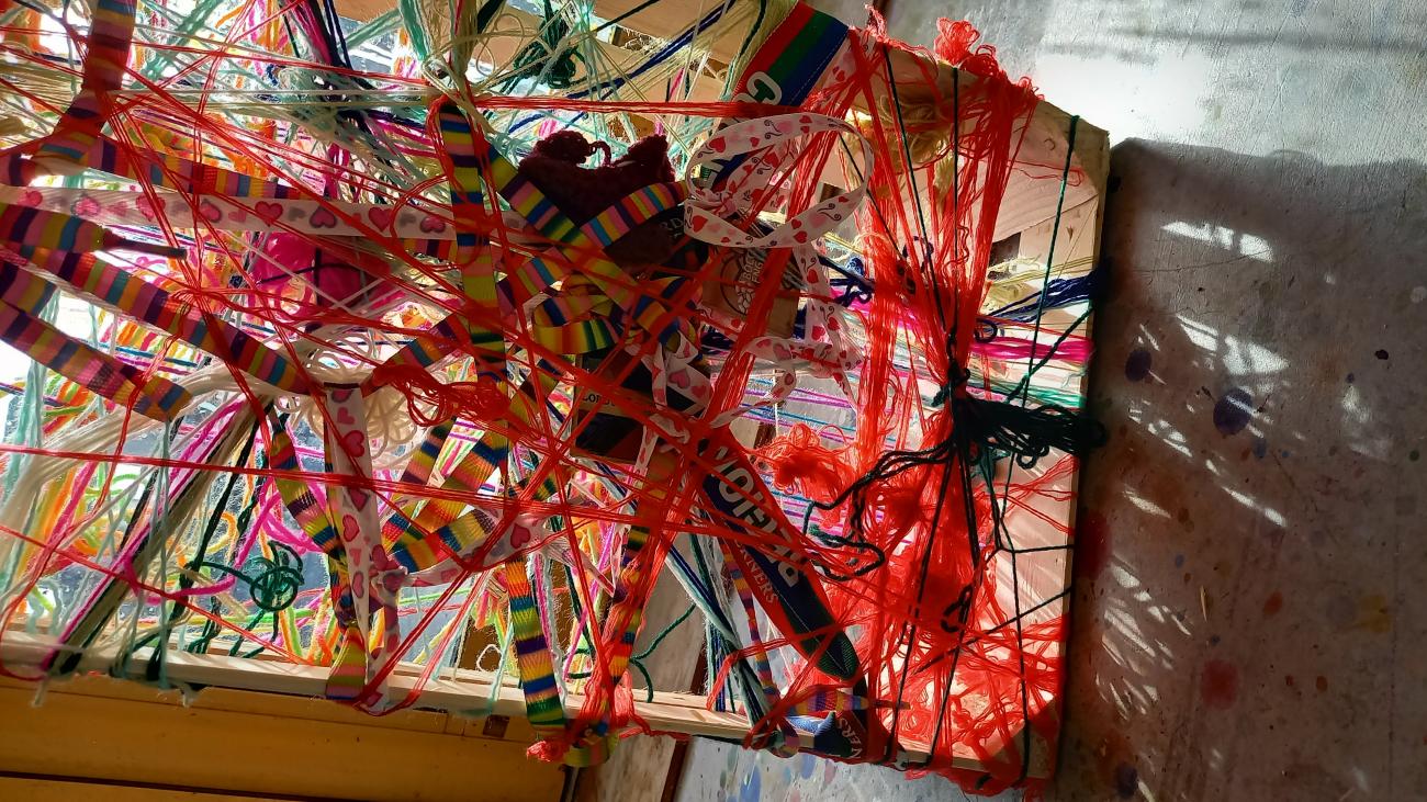 A wooden frame is woven back and forth with brightly coloured wool, laces and threads, making a mesh of colour and texture, lit by strong sunlight from behind.