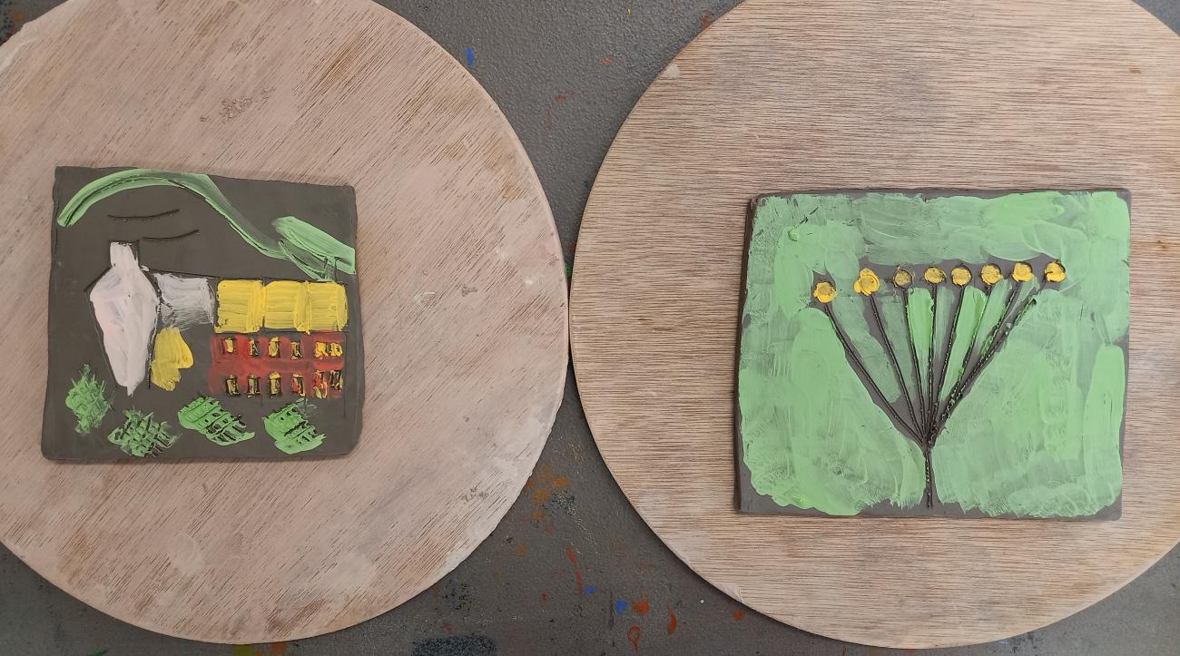 Two flat clay tiles sit on round wooden trays. One clay tile shows a house painted in yellow, red and white. The other shows a seed pod with lots of yellow seeds on a green background.