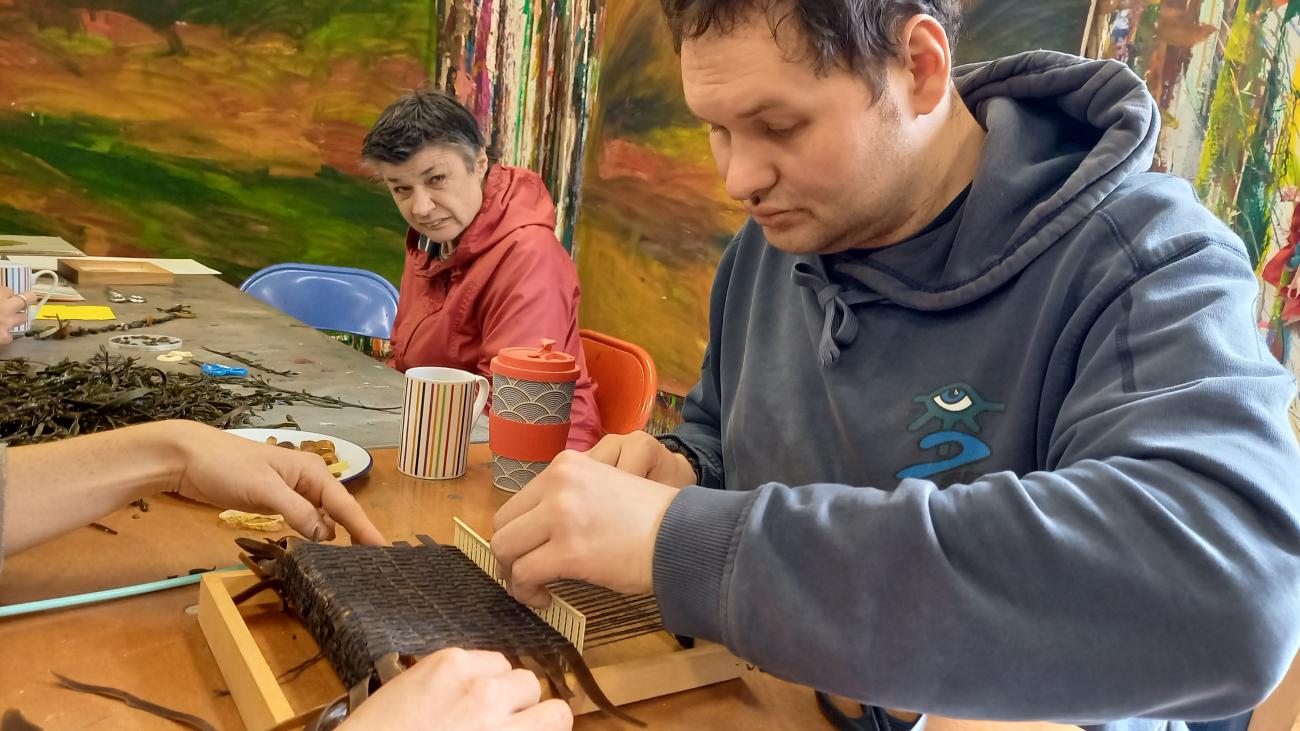 A person is weaving using seaweed and wool on a small loom. The loom is the size of a large book. The person is wearing a grey hoodie and looks interested and happy