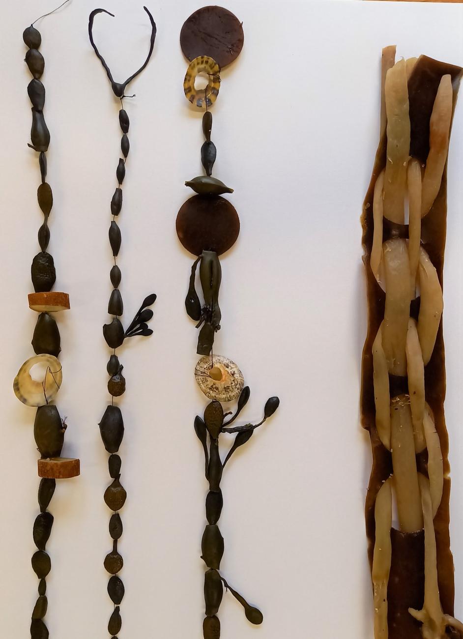 Strings of beads are laid on white paper. They are made from seaweed. They are golden brown with bead-like shapes, shells and shapes like little trees and leafs.