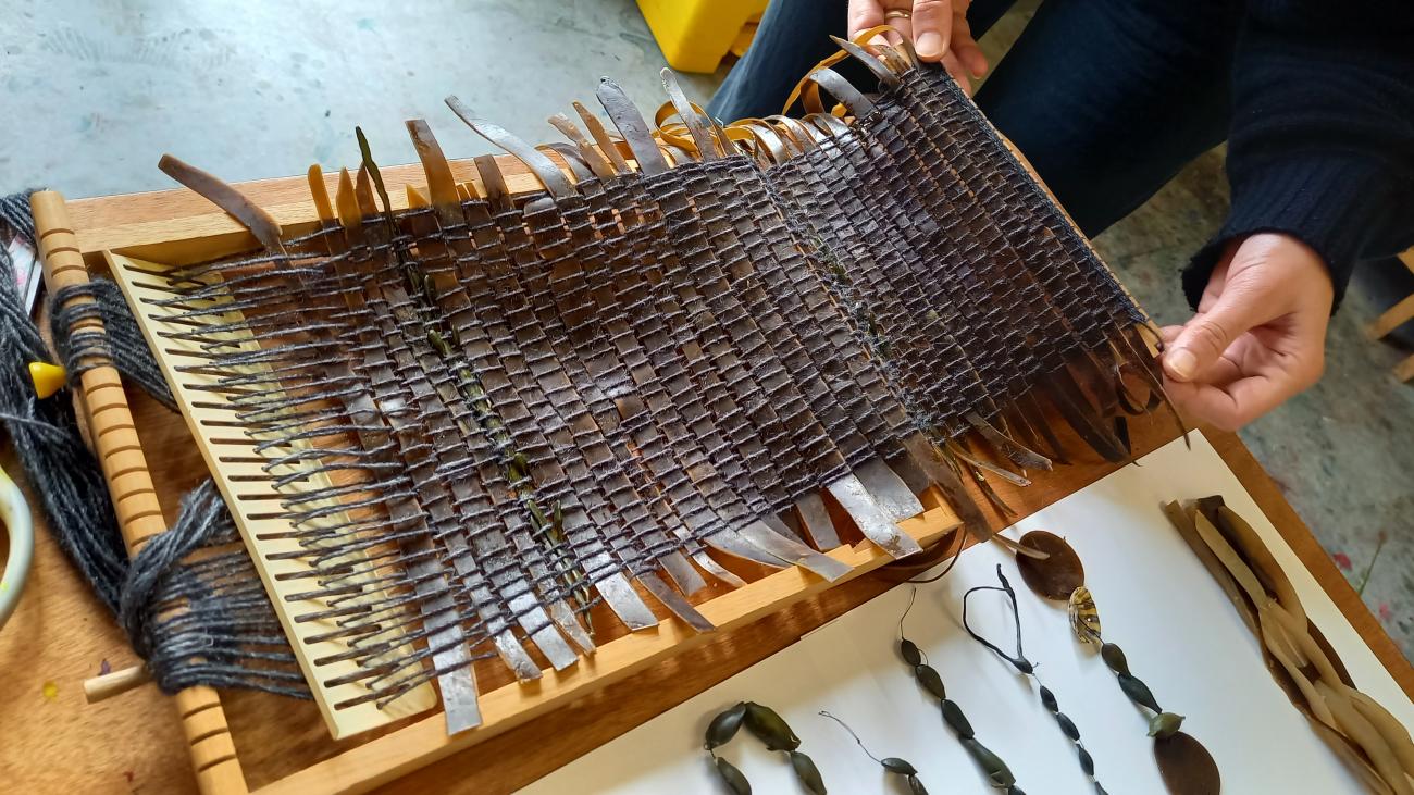 There is a length of brown seaweed strips woven neatly into a flat square in the middle of a small wooden loom. The seaweed is dark and shiny.