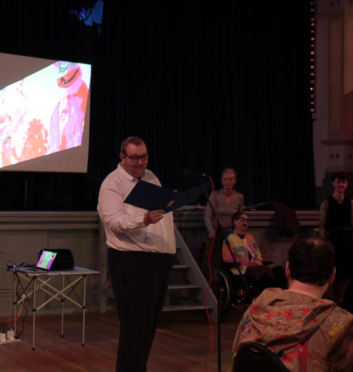 A man wearing a white shirt and black trousers is standing in a dark space with a projector screen behind him. He is talking and people are watching and listening to him.
