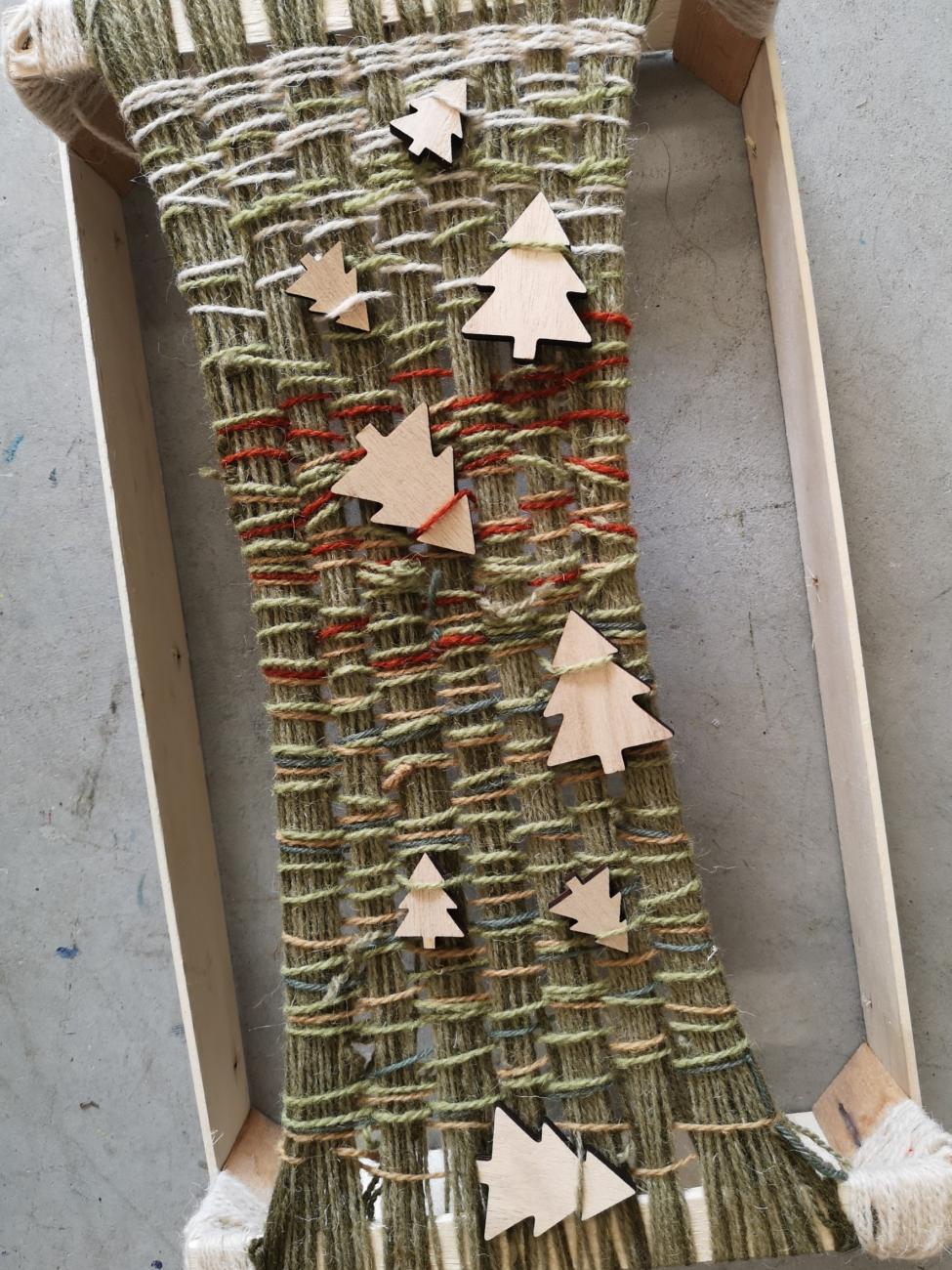 Soft green and brown wool is woven onto a wooden frame, making a chunky, pleasing texture. Little wooden Christmas tree shapes are woven into the piece.