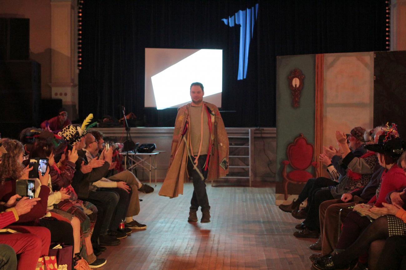 A big brown cape is A tall person is wearing a big cape on the catwalk. The cape is adorned with colourful patterns, images, ribbons and feathers. At the sides we can see people clapping in the audience and images on a projector screen at the back..