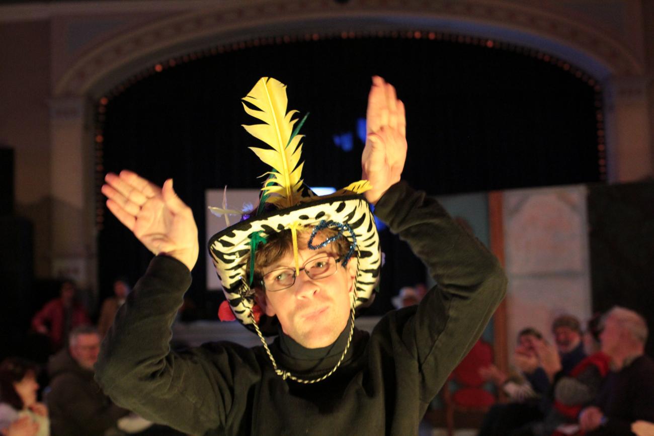 The photo shows a person's head and shoulders. He is wearing a black and white print cowboy hat with a giant yellow feather sticking up from the middle of the brim. He has both his hands up either side of the hat. He is wearing glasses and a black top and is smiling. 