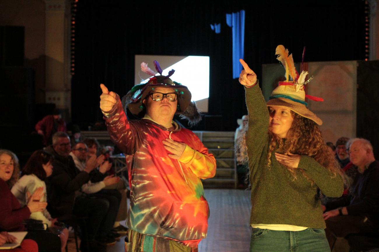 Two people are on the fashion show catwalk. They are both pointing with their right hands and wearing hats with feathers in.